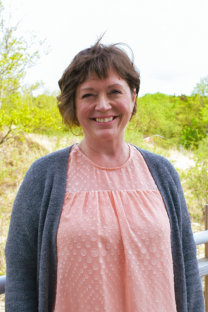 A smiling woman with short brown hair standing outdoors with green trees and a cloudy sky in the background.