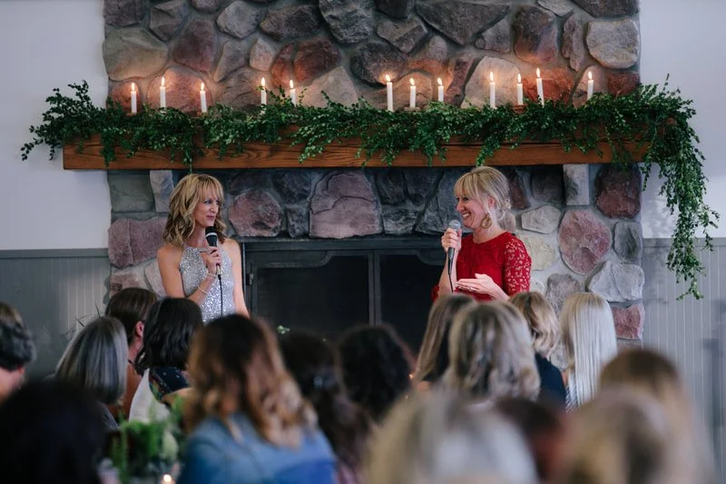 Two women speaking at a gathering in front of a fireplace decorated with candles and greenery; audience seated facing them.