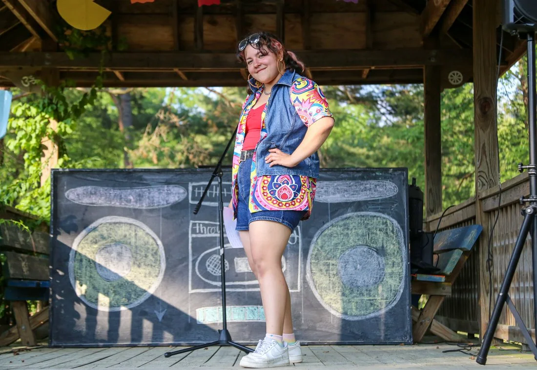 A young girl with curly hair, wearing sunglasses, a colorful dress with an abstract pattern, a denim vest, and white sneakers, stands on a wooden stage with trees in the background. She is smiling with one hand on her hip, in front of a blackboard wi