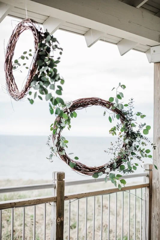 Two decorative wreaths made of twigs, adorned with green eucalyptus leaves, hanging from the ceiling of a porch overlooking the beach.