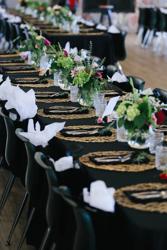 Set dining table with black tablecloths, gold placemats, white napkins, glassware, and floral centerpieces.