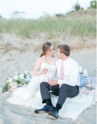 Couple sitting on the beach during a wedding ceremony, surrounded by flowers and greenery.