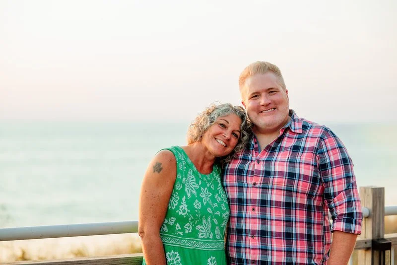A smiling woman with gray curly hair and a tattoo on her arm, wearing a green dress, standing next to a smiling man in a red and blue plaid shirt, both posing outdoors on a boardwalk near the ocean at sunset.