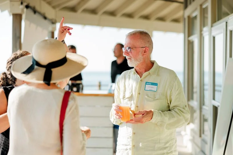 People socializing on a balcony with ocean view, a man with glasses holding a drink, woman with a large sun hat, other people in the background.