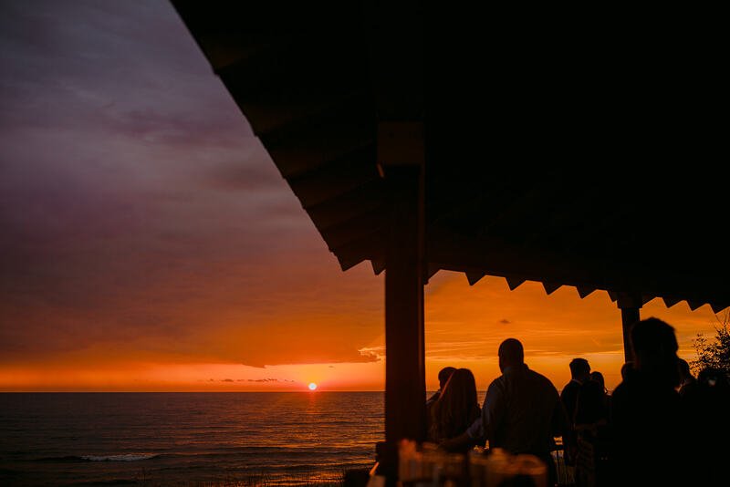 Silhouettes of people standing under a shaded structure, watching a sunset over the ocean.