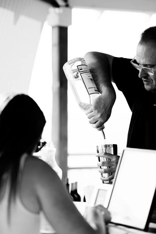 A man pouring a drink into a glass at a bar or restaurant, with a woman sitting in front of him, seen in a black-and-white photo.