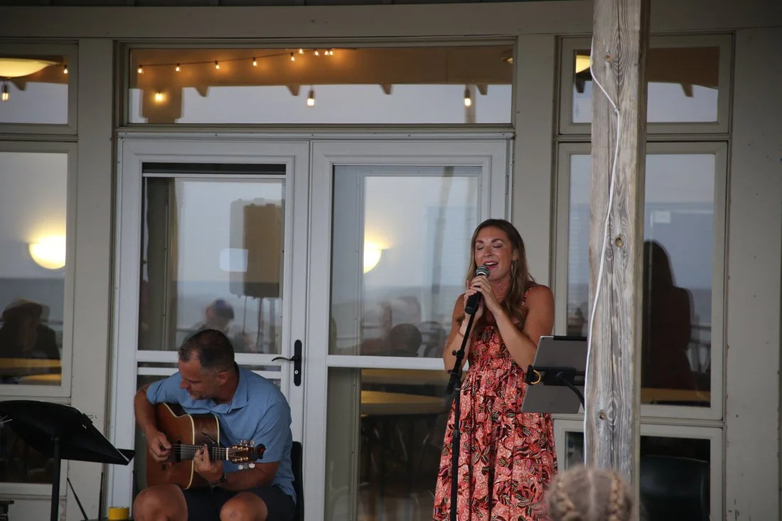 A woman singing into a microphone and a man playing guitar at an outdoor event with a glass door and string lights in the background.