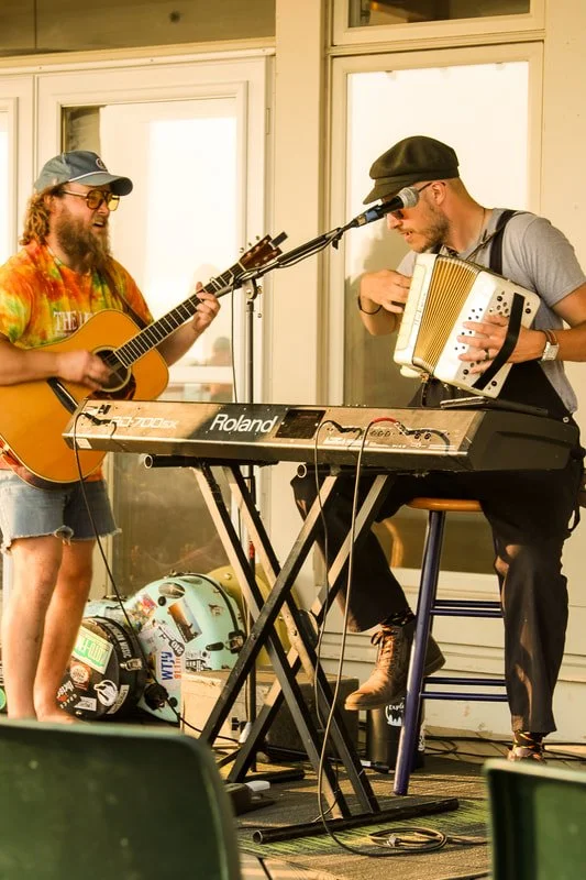 Two musicians performing together on a balcony with large windows, one playing an acoustic guitar wearing a colorful shirt and shorts, the other playing an accordion and sitting on a stool wearing a hat and glasses.