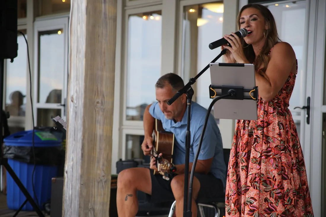 A woman singing into a microphone with a tablet in front of her, and a man playing guitar seated behind her.