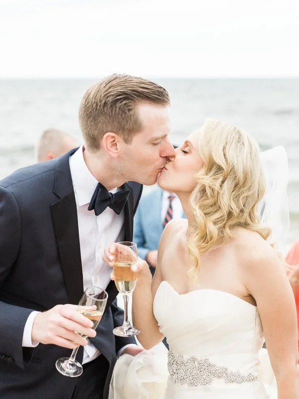 A newlywed couple sharing a kiss at a beach wedding celebration, holding glasses of champagne.