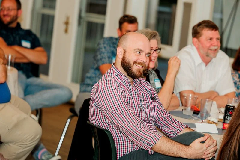 Group of people sitting around a table at an indoor gathering, smiling and engaging with each other.