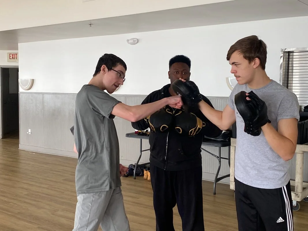Two young men practicing boxing with boxing gloves while an instructor observes.