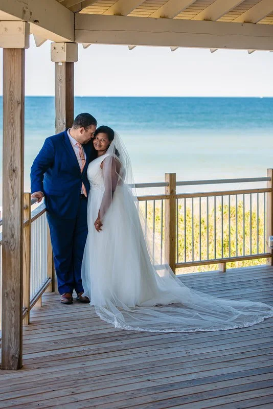 A bride and groom standing on a wooden deck by the ocean, dressed in wedding attire, with the groom in a blue suit and the bride in a white gown with a veil.