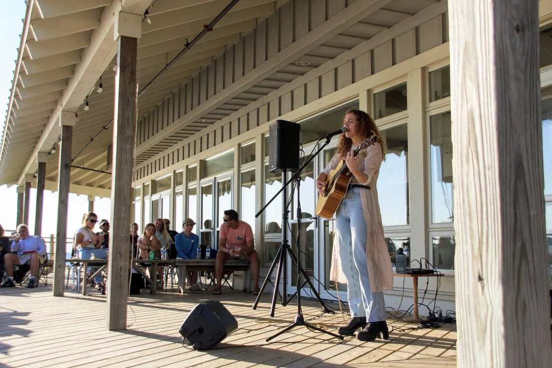 A woman playing guitar and singing on an outdoor wooden deck with a small audience watching.