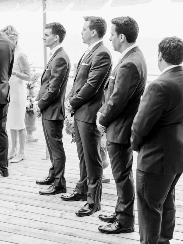 A line of men in suits at a formal outdoor event, possibly a wedding, standing on a wooden deck.
