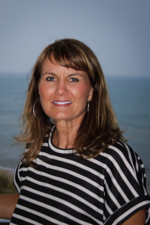 A woman with shoulder-length brown hair wearing a black and white striped shirt, standing outdoors near the water with a blue sky background.