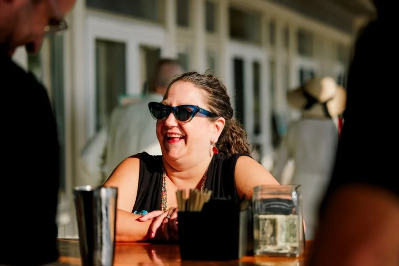 A woman with curly hair wearing sunglasses and red earrings smiling at a bar or outdoor restaurant.