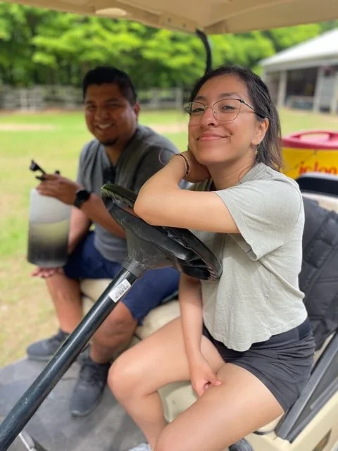 A young woman sitting on a golf cart with her arm resting on the steering wheel, smiling, with a young man sitting behind her holding a drink, outdoors on a golf course.
