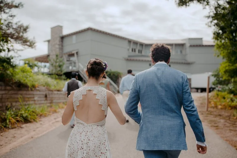 A couple walking away from the camera, with the woman wearing a white lace dress and the man in a blue suit, towards a large house in the background.