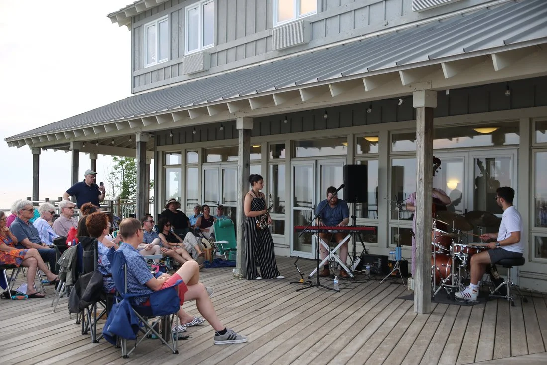 Musicians performing on a wooden deck in front of an audience at an outdoor event, with a house or restaurant in the background.