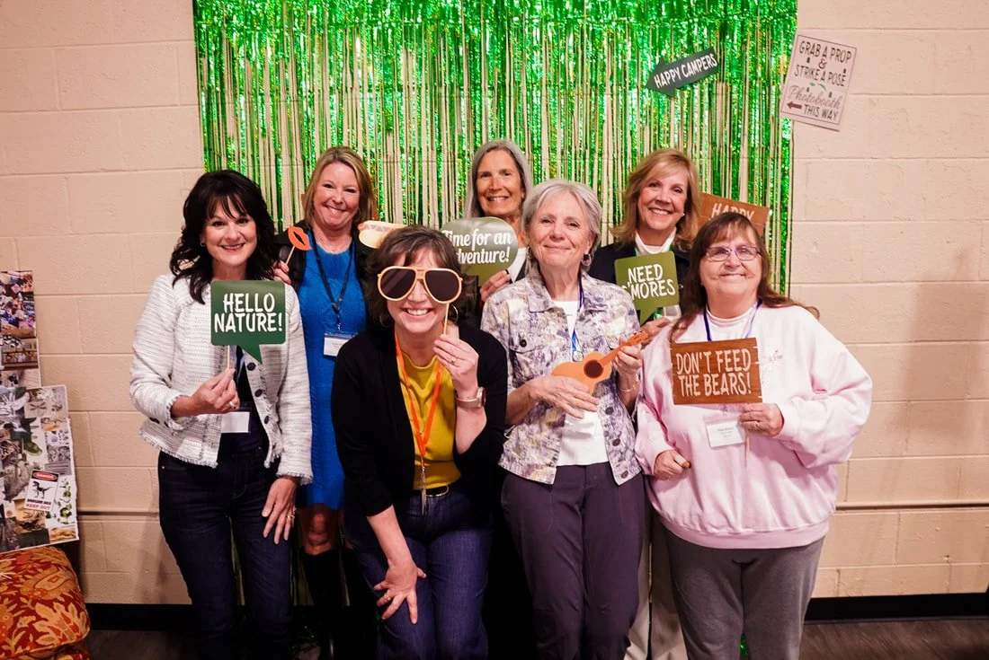 A group of eight women posing together at a celebration with a green tinsel backdrop. They are holding signs with outdoor themes and wearing fun props like large sunglasses. The signs read "Hello Nature!", "Time for an Adventure!", "Need Mores", and 