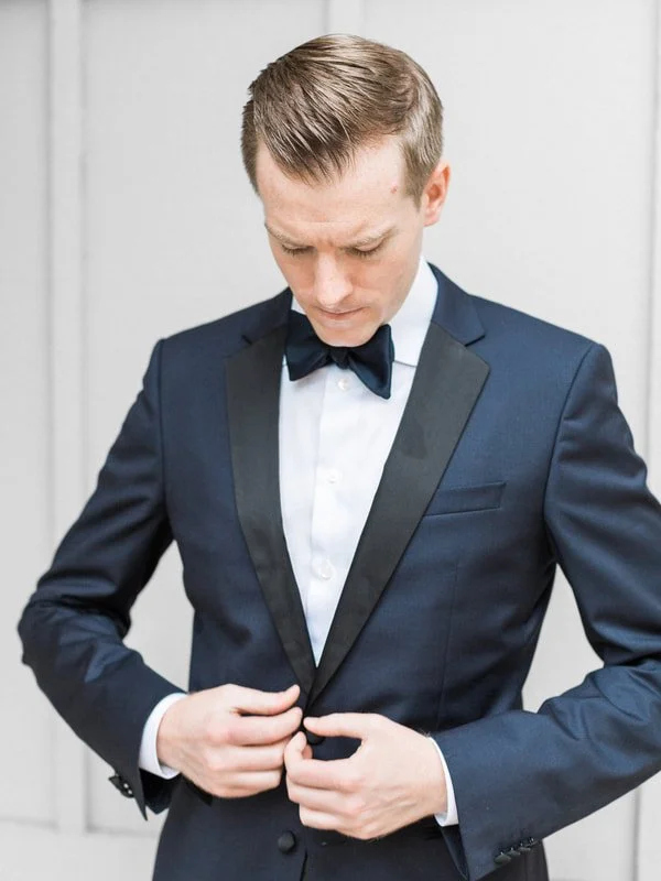 A young man in a dark blue tuxedo with a black lapel, white dress shirt, and black bow tie buttoning his jacket.