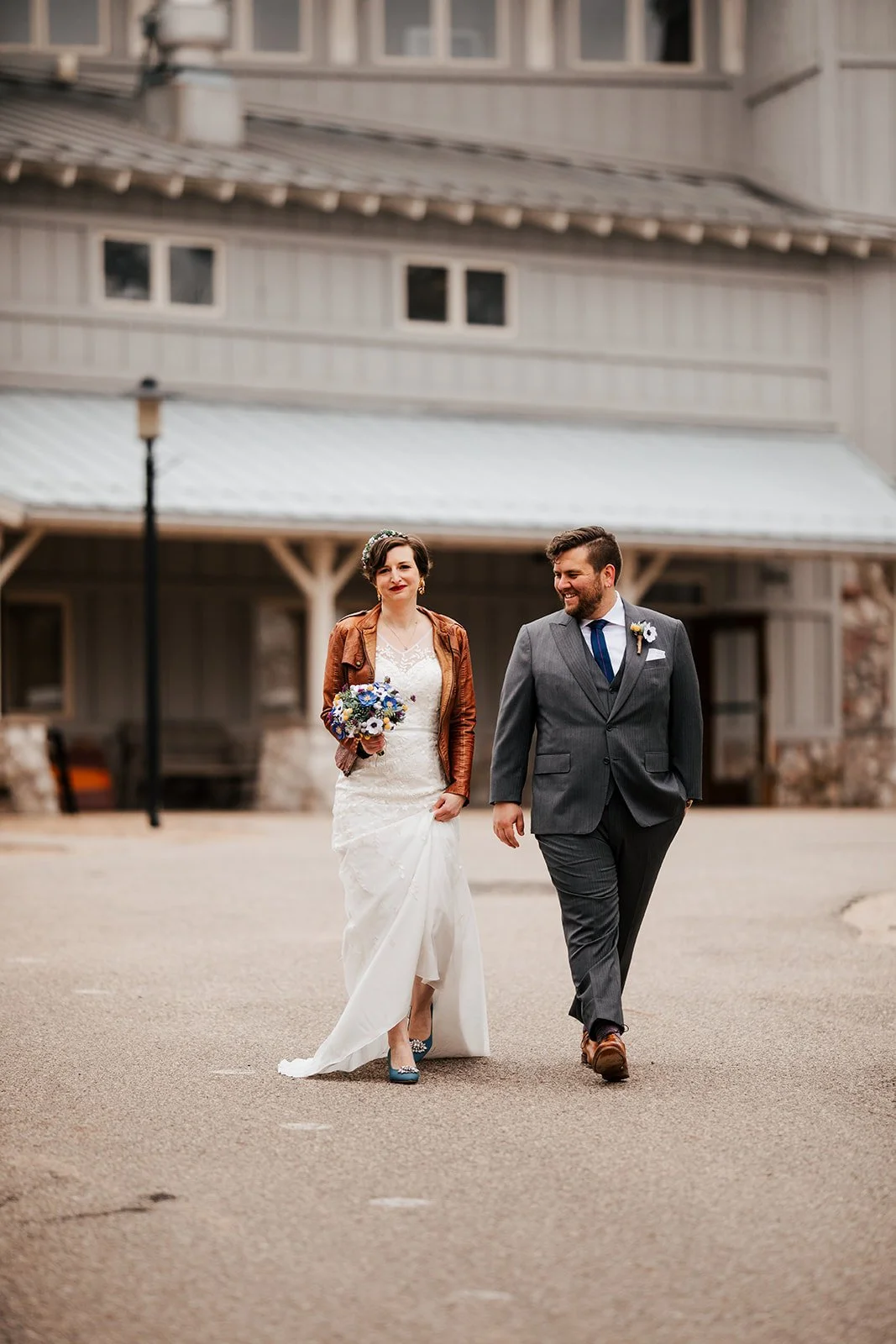 A couple walking outdoors in wedding attire, with the bride holding a bouquet and the groom smiling, in front of a rustic building.