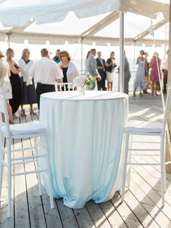 A decorated table with a white tablecloth and floral centerpiece under a white canopy on a deck, with blurred people in the background at an outdoor event.