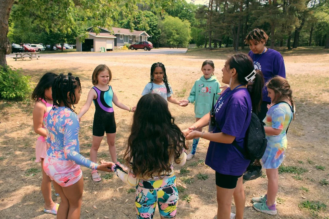 A group of children holding hands in a circle outdoors, with two adults supervising, in a park with trees, grass, and a building and cars in the background.