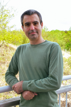 A man with short dark hair wearing a green long-sleeve shirt, standing outdoors by a railing with greenery in the background.
