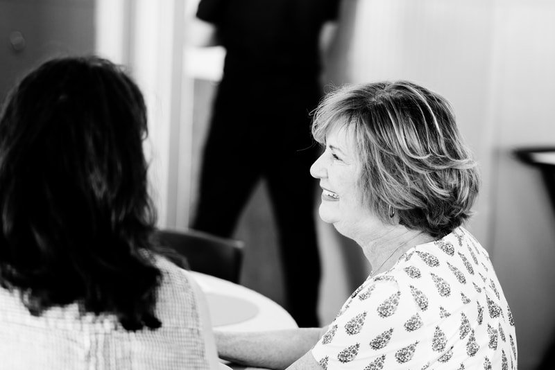 An elderly woman with short, wavy hair smiling while talking to a person with dark hair in a social setting, possibly a conversation at a gathering.