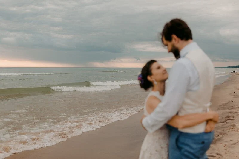 A couple embracing on a beach during sunset, with gentle waves and a cloudy sky in the background.
