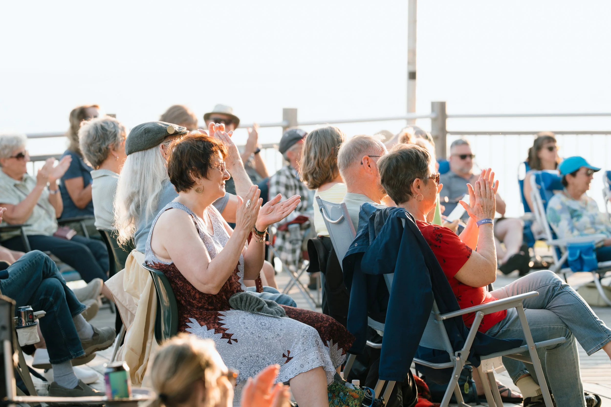 Group of people sitting on chairs outdoors, enjoying a performance or event, some clapping, some wearing sunglasses.