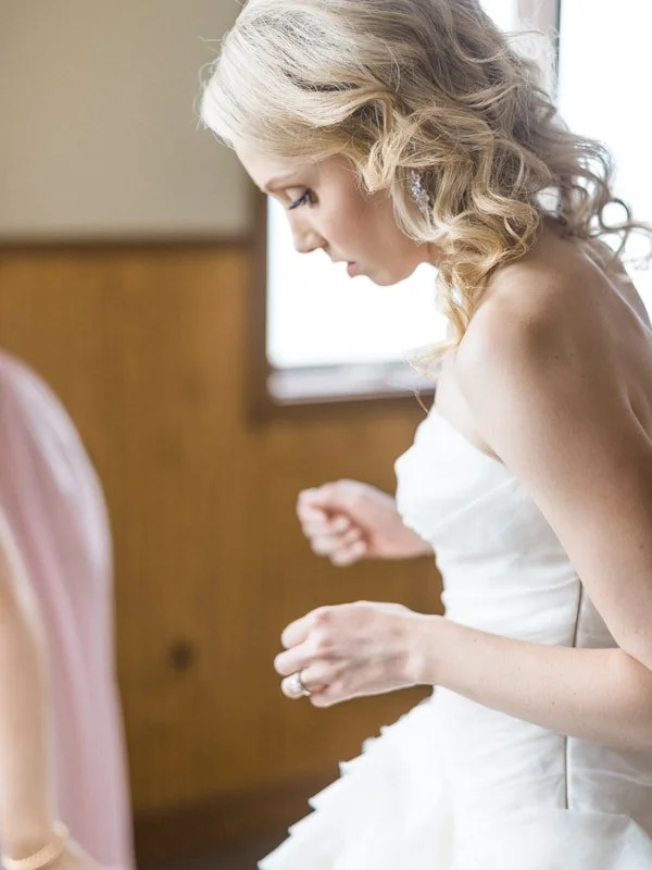 A bride with blonde curly hair wearing a white strapless wedding dress, standing indoors with a wooden-paneled wall and a window in the background, adjusting her ring.