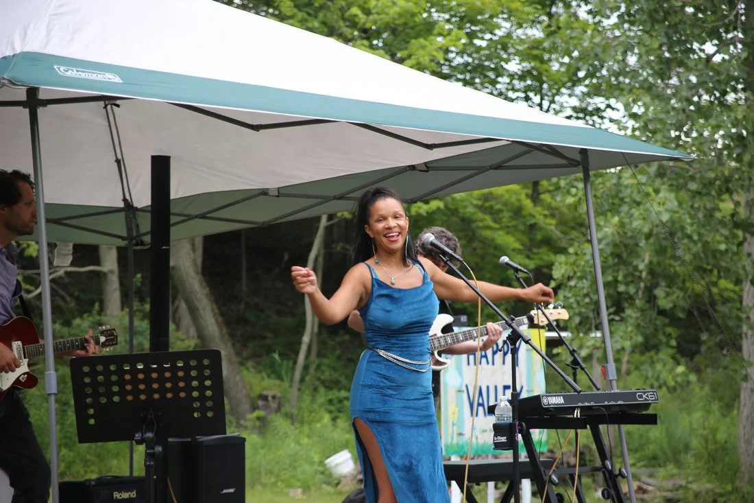 A woman singing and dancing on an outdoor stage with musicians, under a canopy, with green trees in the background.