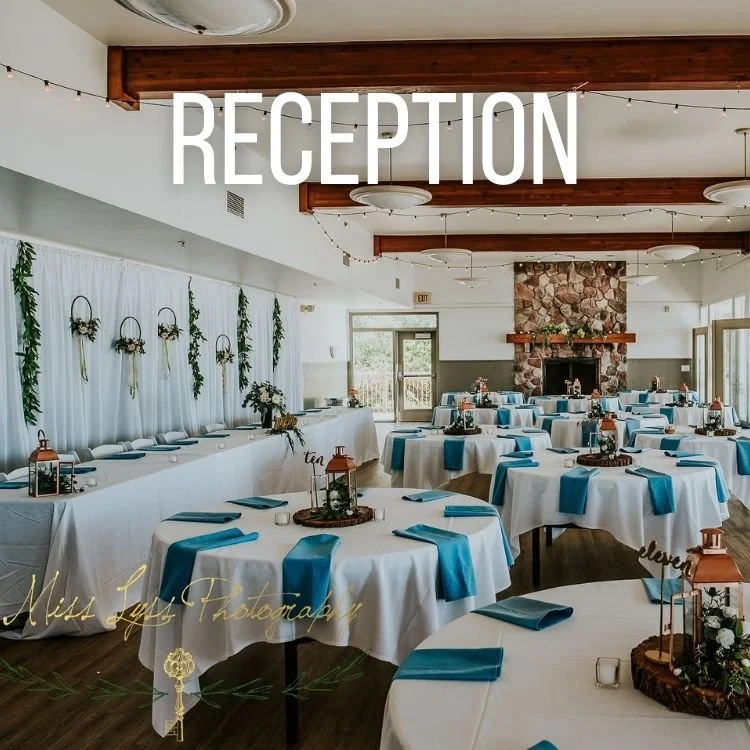 Wedding reception hall decorated with white curtains, green foliage, and string lights. Round tables with white tablecloths and blue napkins. Centerpieces feature lanterns, candles, and floral arrangements. Fireplace with a stone surround is at the back of the room.
