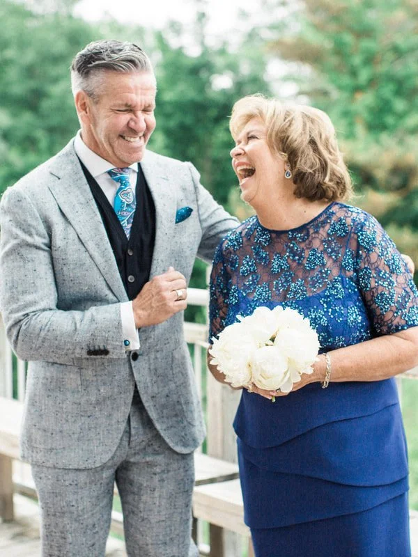 A man and a woman sharing a joyful moment outdoors, with the woman holding a bouquet of white flowers, both smiling and laughing.