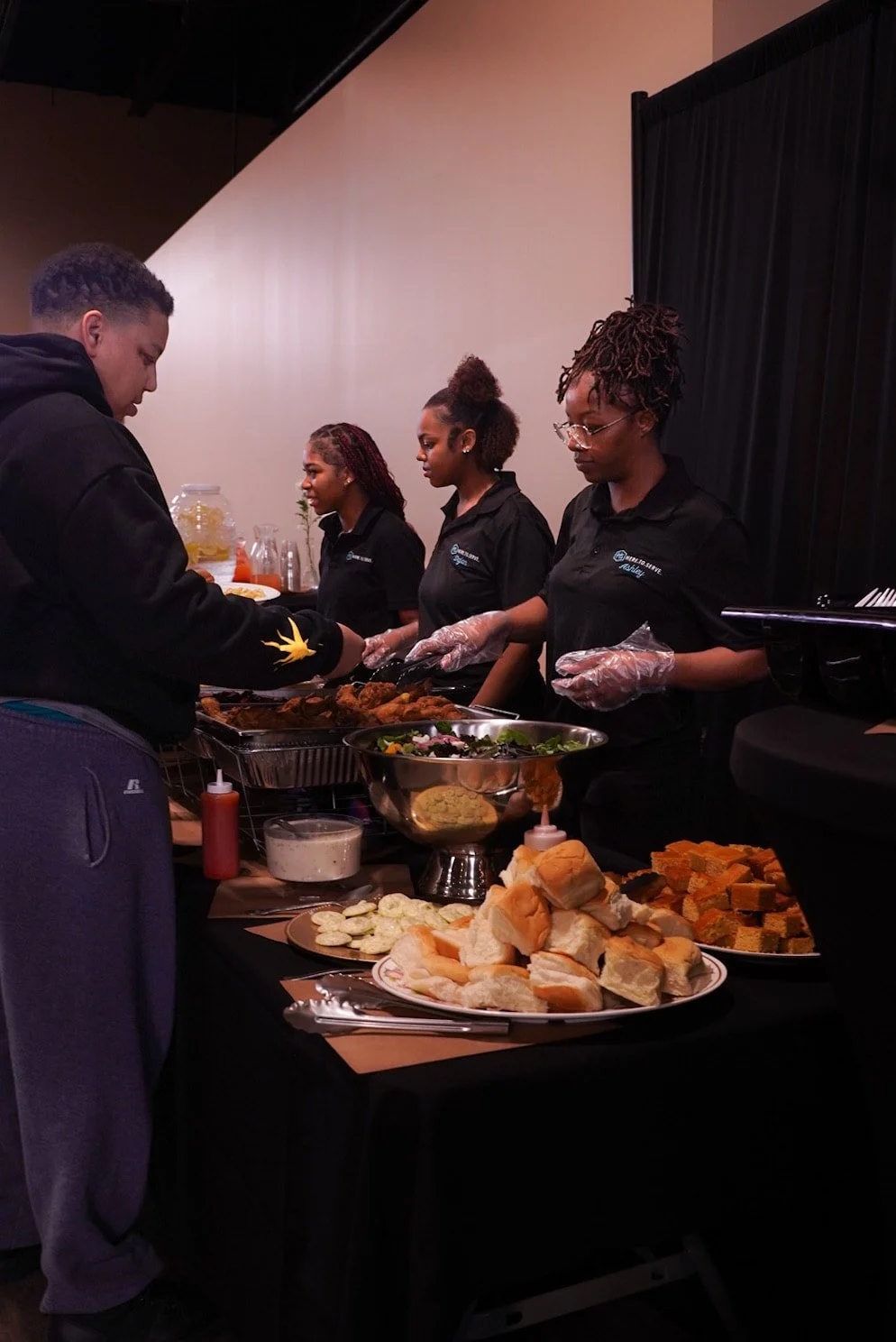People serving food at a buffet-style table with various dishes and drinks.
