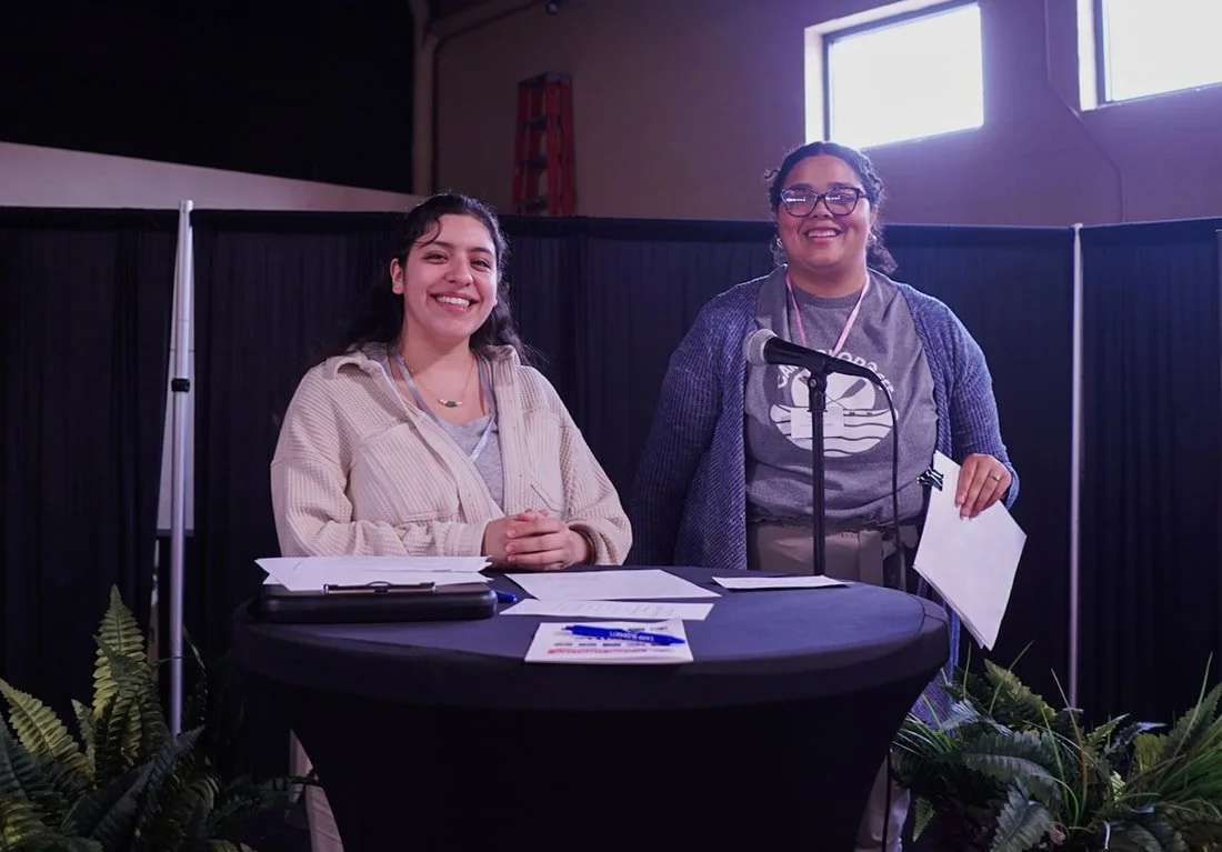 Two women smiling at a table with papers, in front of a microphone, inside a room with large windows and black curtains.