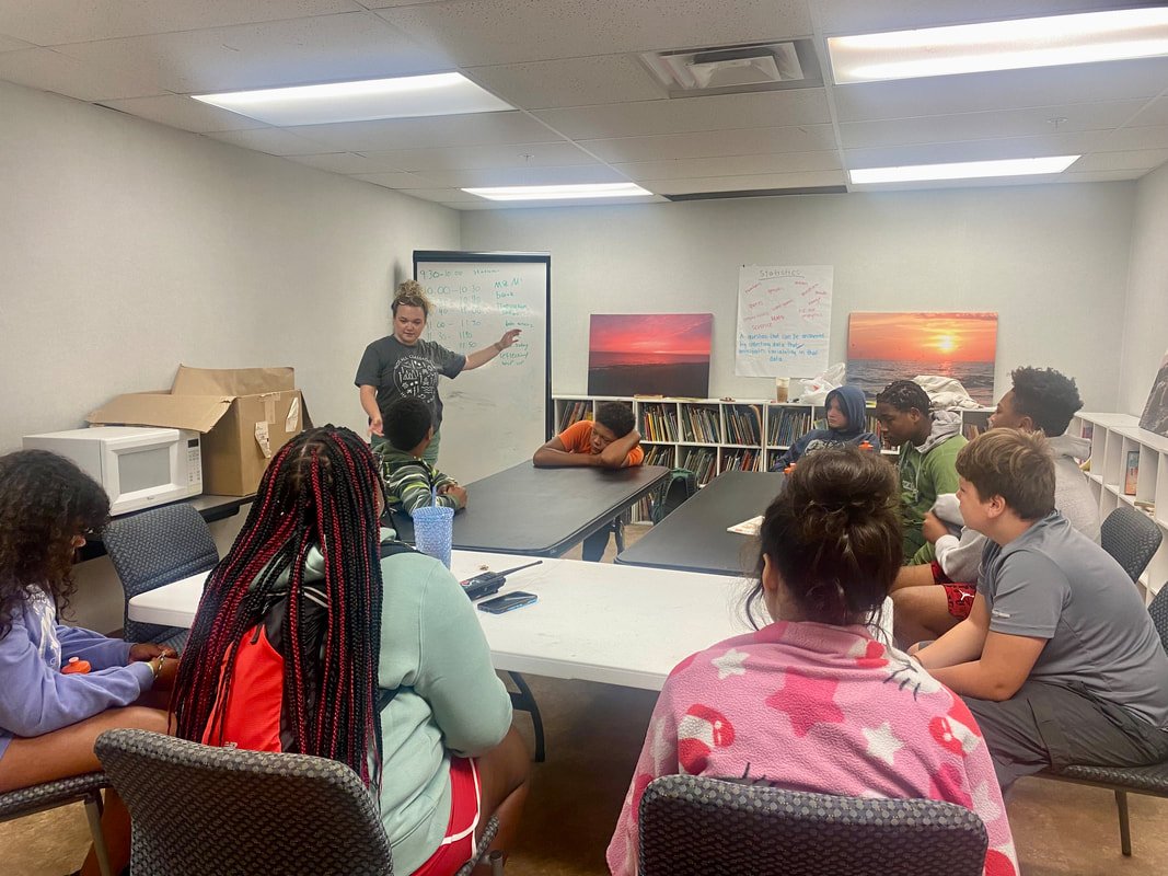 A classroom with a group of students sitting around a large conference table while a woman appears to be leading a discussion or lesson at the whiteboard.