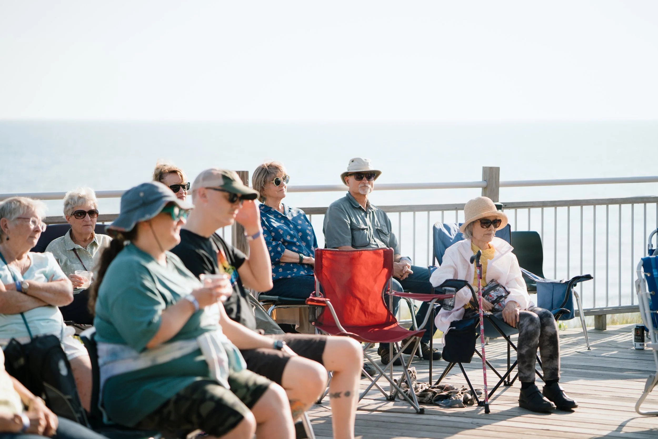 Group of elderly people sitting in outdoor chairs on a wooden deck near the ocean, some wearing hats and sunglasses, enjoying a sunny day.