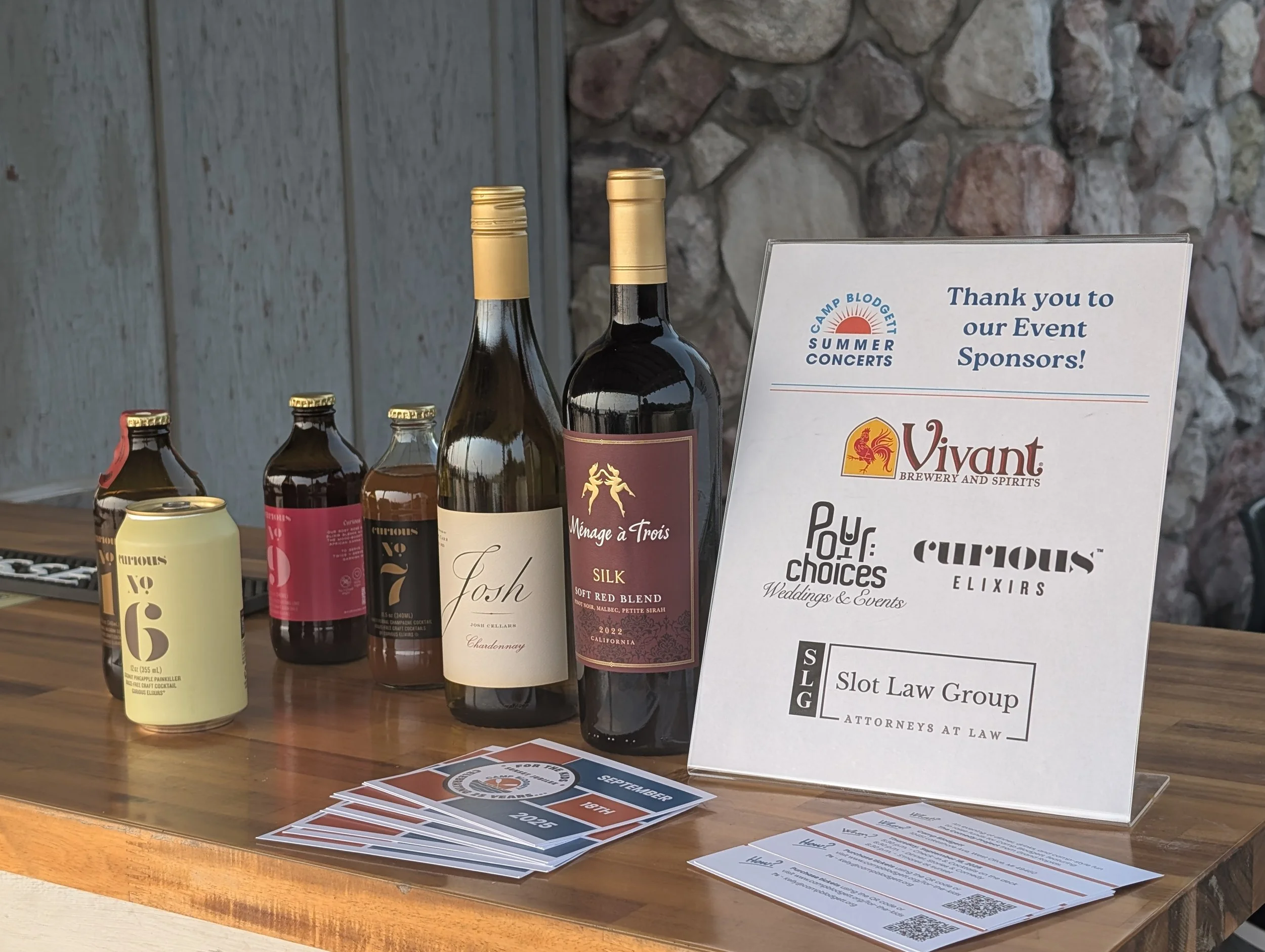 A wooden table displaying six bottles of beverages, a can, and informational flyers, with a sign thanking event sponsors, including Vivant Brewery and Spirits, Pop Choices Weddings & Events, Curious Elixirs, and Slot Law Group, with a stone wall in t