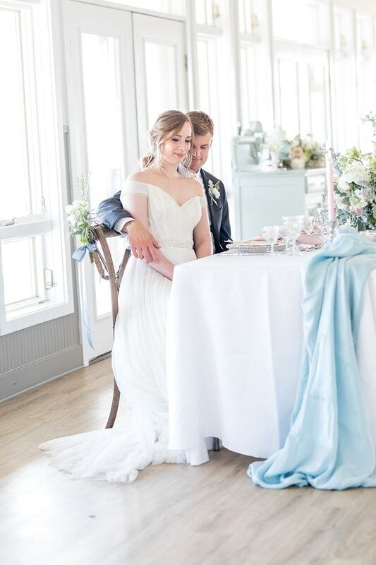 A bride wearing a white wedding gown and a groom in a dark suit stand together at a wedding reception table in a bright, airy room with large windows and floral decorations.
