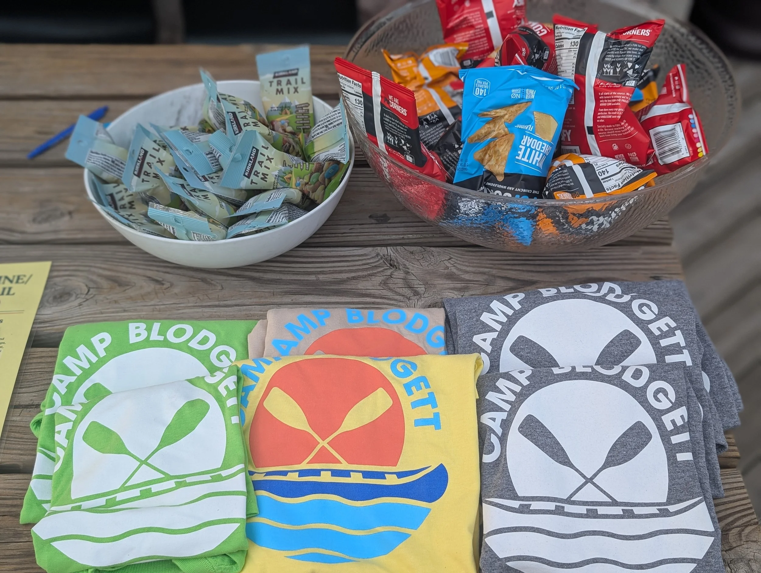A wooden table with two bowls filled with snack-sized packages, one containing trail mix and the other chips. Below the bowls are folded T-shirts with the text "CAMP BLODG" and a graphic of crossed paddles, in green, blue, yellow, and gray colors.