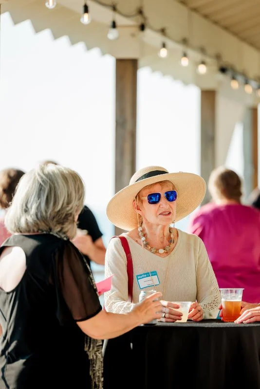 A senior woman wearing a wide-brimmed sunhat, sunglasses, and a beaded necklace talking to another woman at a social event with drinks and name tags.