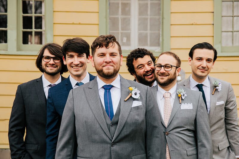 Six men dressed in suits posing outdoors in front of a yellow house with green trim.