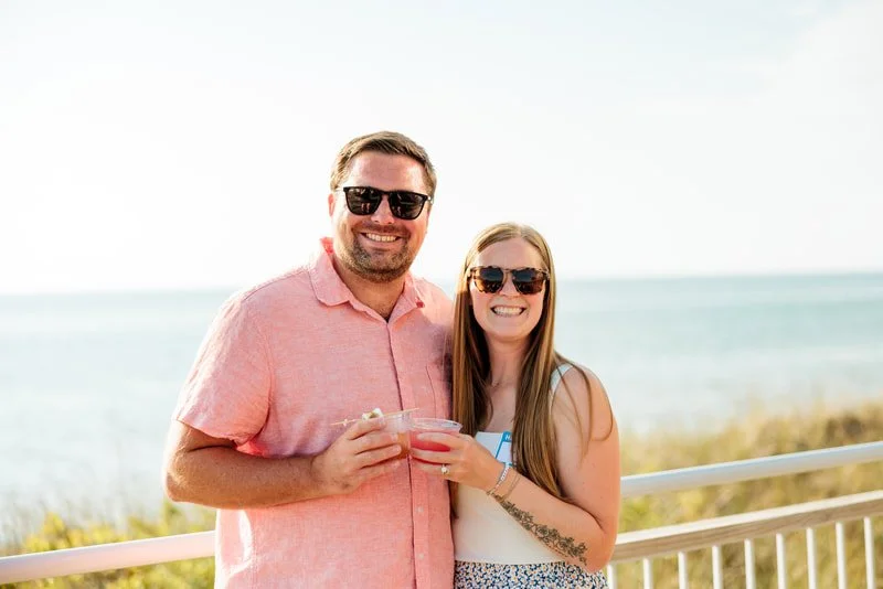 A smiling man and woman wearing sunglasses, holding drinks, standing outdoors near the beach with a railing and ocean in the background.