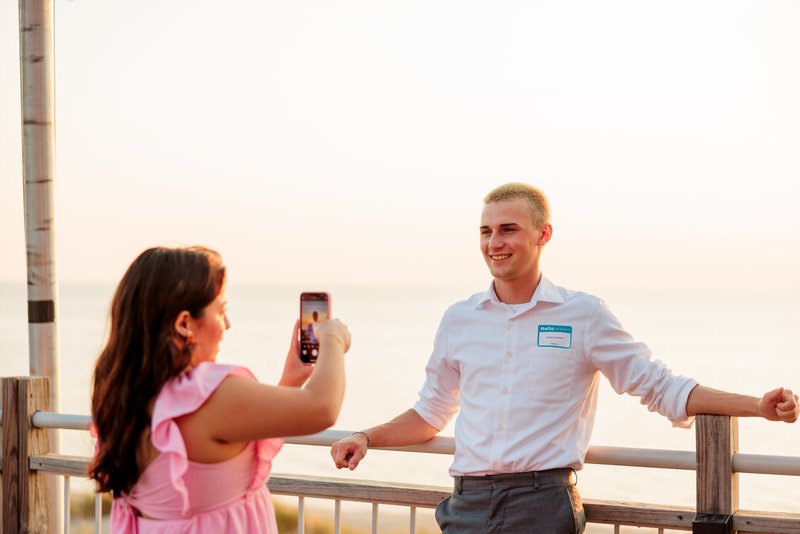 A woman is taking a photo of a man on a balcony overlooking a body of water during sunset.