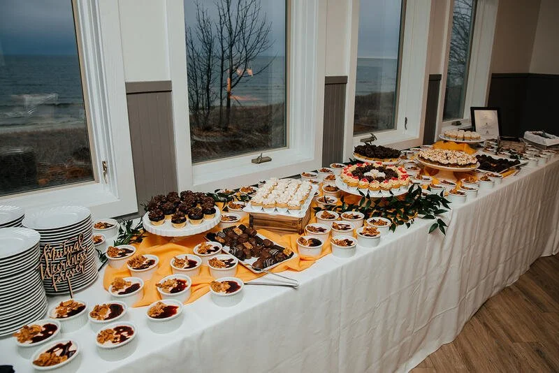 A long table with white tablecloth holding various desserts, including cupcakes, brownies, cakes, and small bowls of fruit or pudding, with large windows overlooking a beach or ocean during sunset in the background.