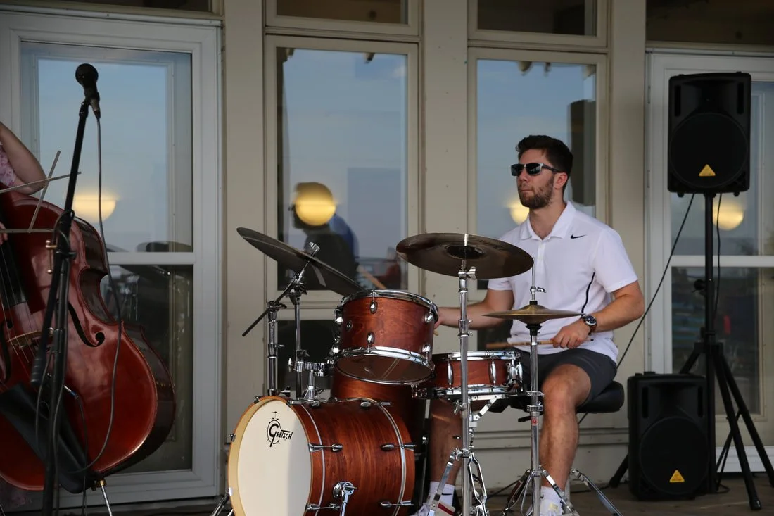 A man wearing sunglasses, a white shirt, and shorts playing a drum set on an outdoor balcony with windows and a speaker.